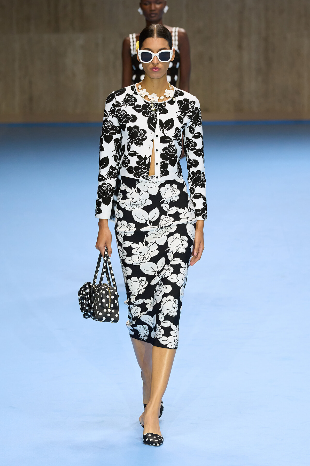 Model walking runway in black-and-white floral suit with oversized white sunglasses and matching polka dot bag and shoes, blue floor backdrop.