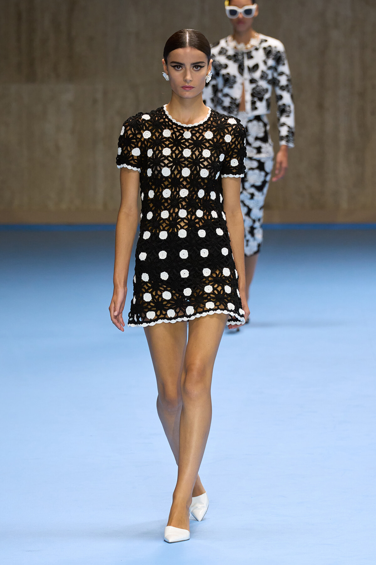 Female model walking a runway in a black crochet mini dress with white circular motifs and white trim, paired with white heels.