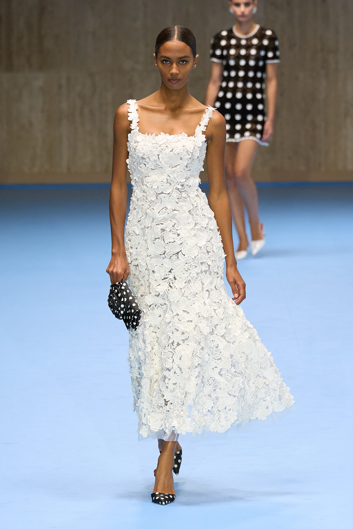Model walking a runway in a white floral lace midi dress, holding a black polka-dot clutch; another model in a black polka-dot dress visible in the background.