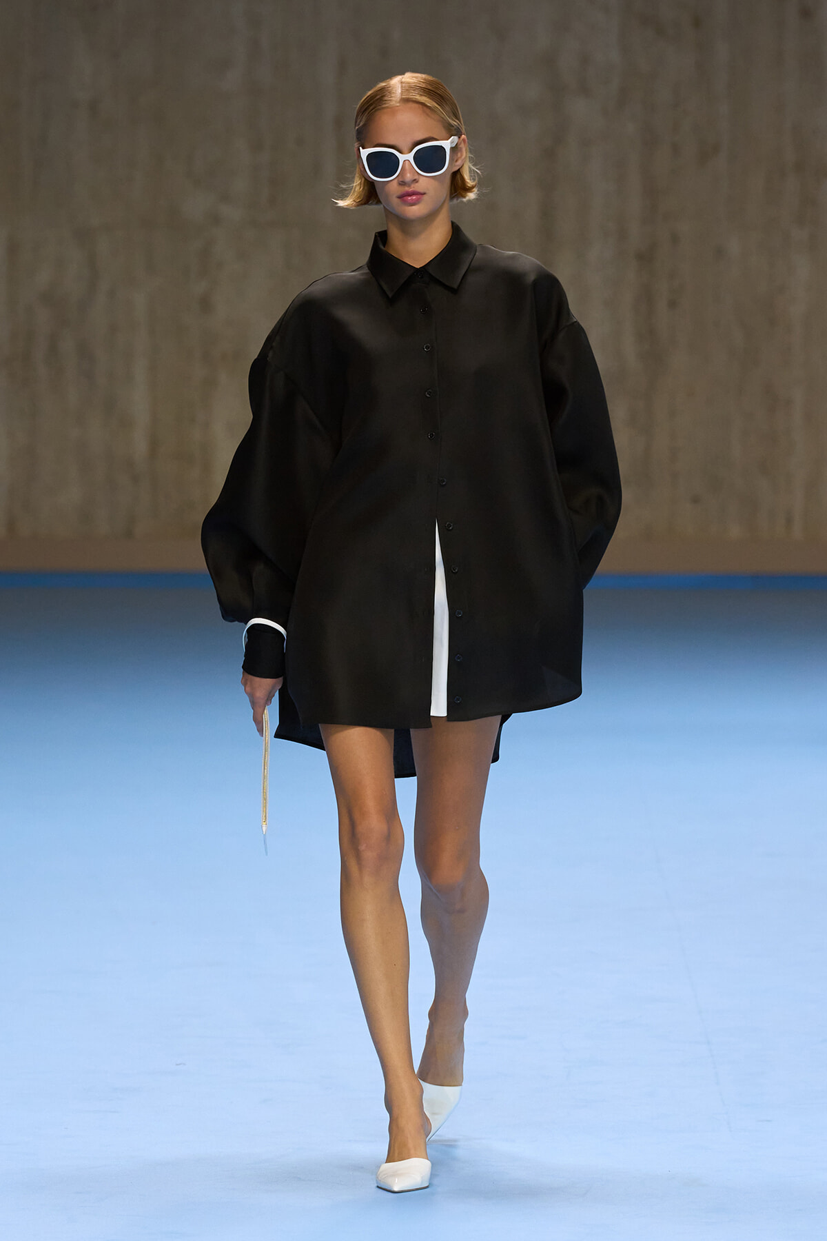 Model on a runway wearing an oversized black button-down shirt dress, white sunglasses, and white pointed heels against a blue floor.