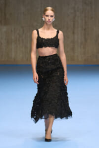 Model walking a runway in a black lace crop top and matching midi skirt with floral appliqués.