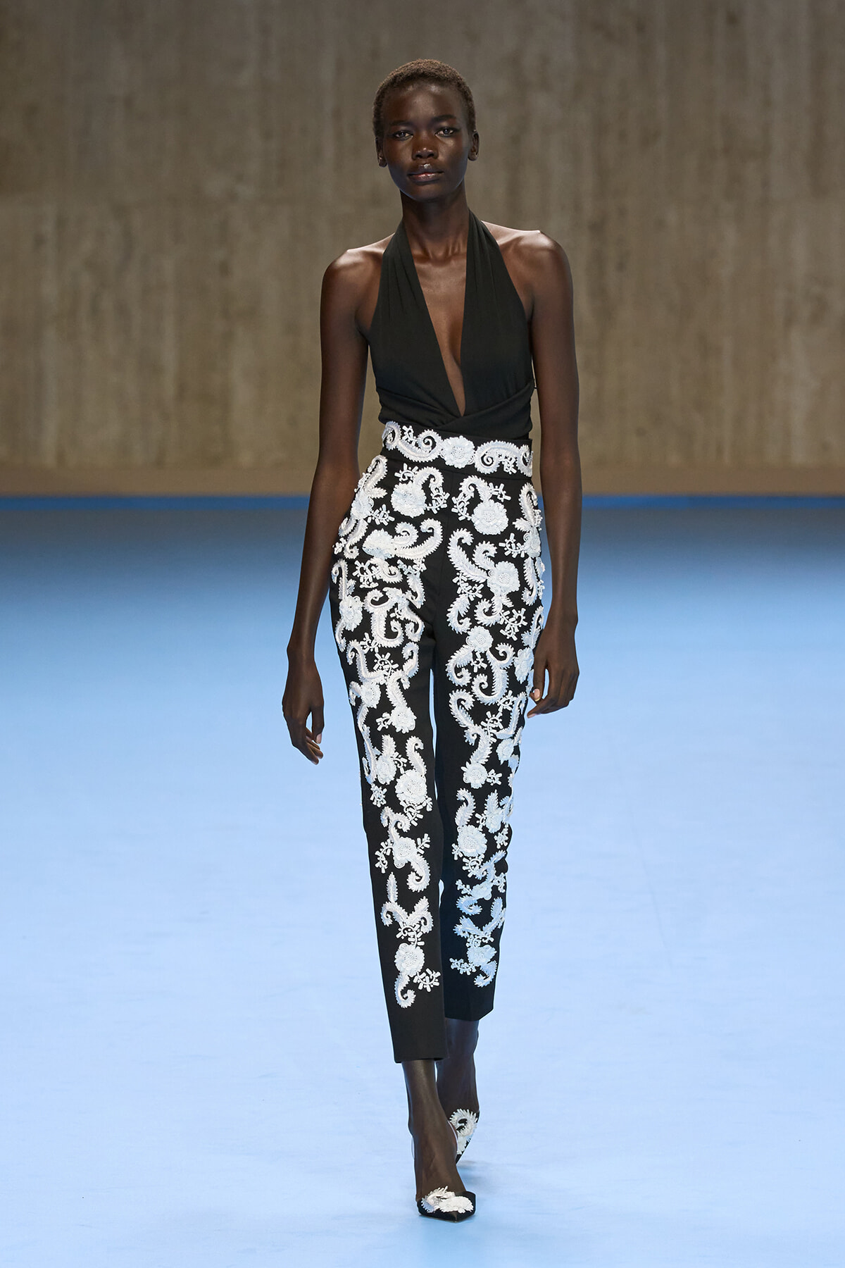Model on a runway wearing a black halter top and black pants with white ornate embroidery.