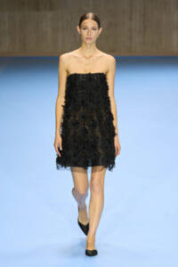Model wearing a strapless black floral-embellished mini dress walking on a blue runway.