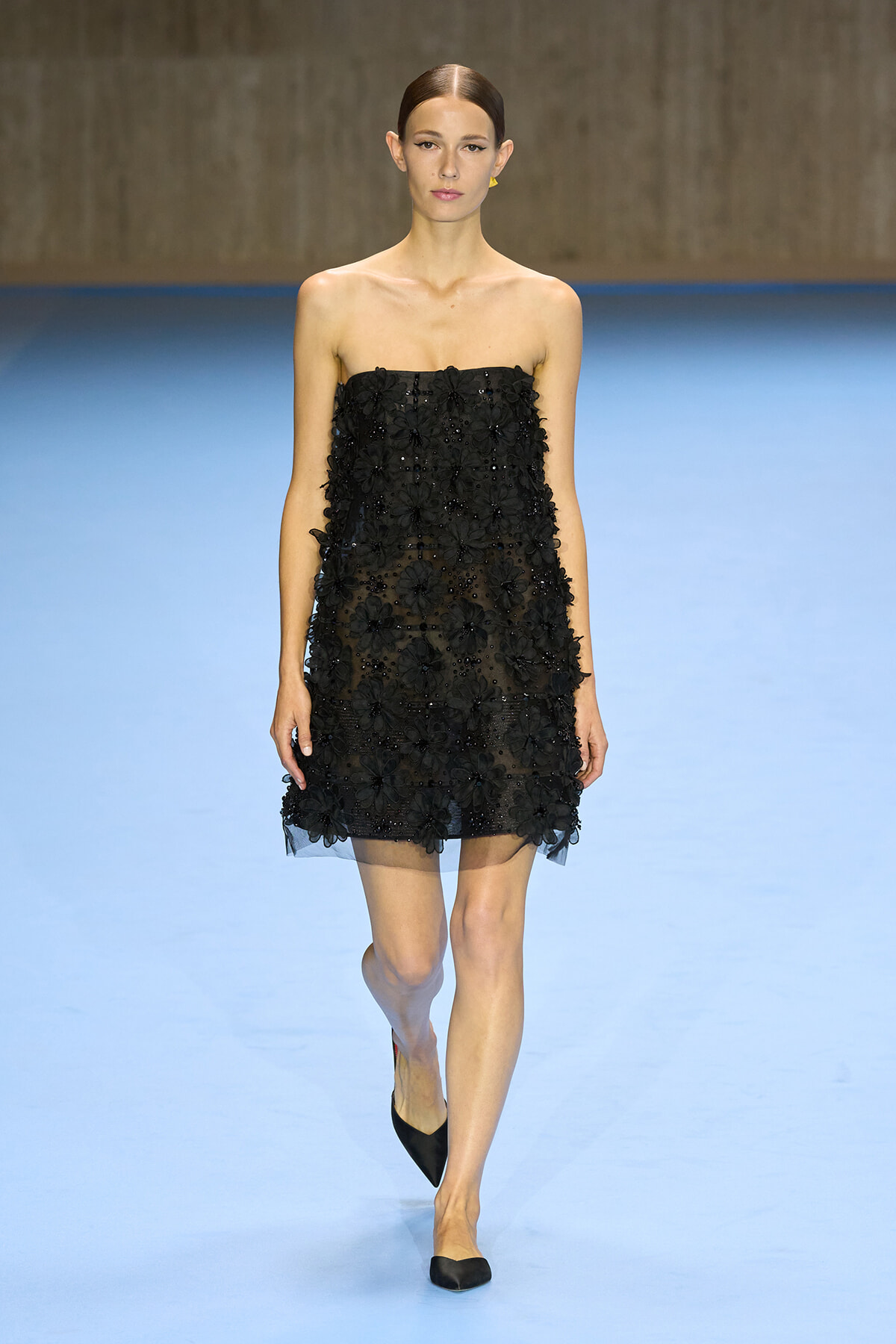 Model wearing a strapless black floral-embellished mini dress walking on a blue runway.