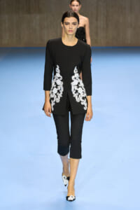 Female model on a fashion show runway wearing a black tailored coat with white floral embroidery along the front sides, paired with black cropped pants and embroidered flats.