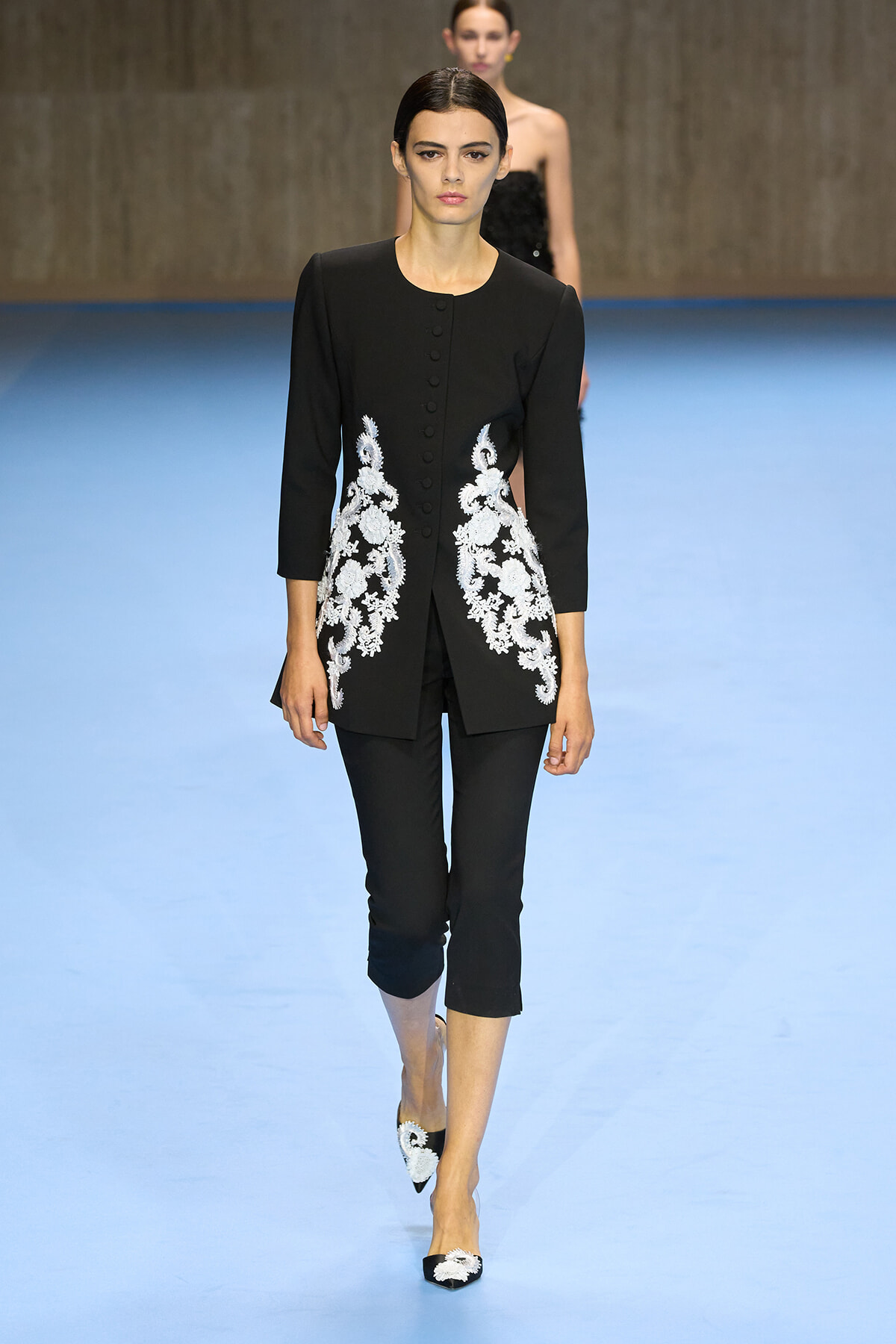 Female model on a fashion show runway wearing a black tailored coat with white floral embroidery along the front sides, paired with black cropped pants and embroidered flats.