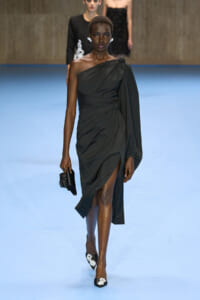 Model on a blue runway wearing a one-shoulder black draped gown with a waist knot and high slit, carrying a black clutch.