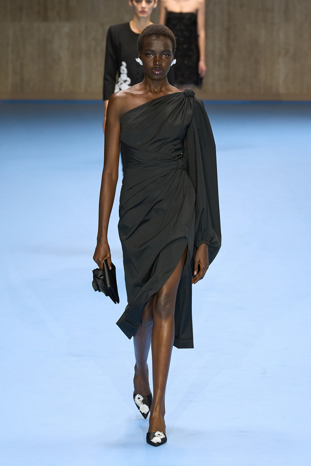 Model on a blue runway wearing a one-shoulder black draped gown with a waist knot and high slit, carrying a black clutch.