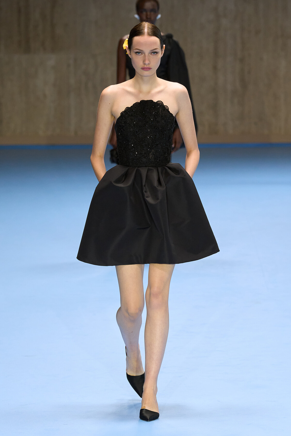 Model walking the runway in a strapless black sequin bodice and full satin mini skirt on a blue catwalk.