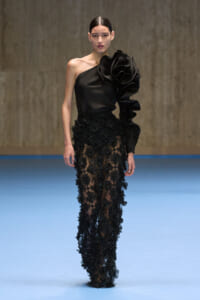 Model on a runway wearing a one-shoulder black satin top with a large ruffle and sheer floral lace pants.