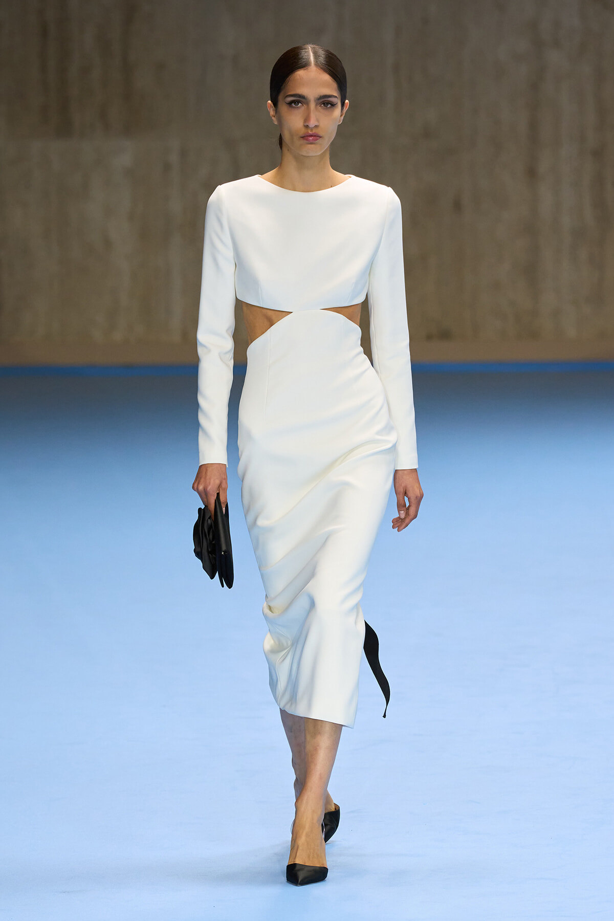 Model walking the runway in a white long-sleeve two-piece dress with a waist cutout, carrying a black clutch and wearing black heels on a blue runway.