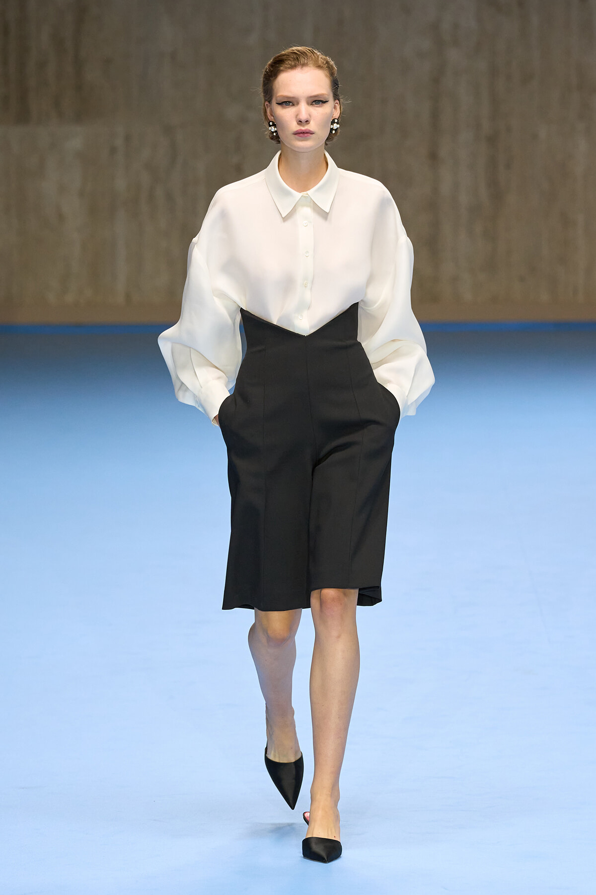 Model walking a runway in a white button-up blouse with oversized sleeves and a black high-waisted knee-length skirt, black pointed heels.