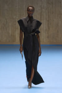 Model wearing a structured black dress with oversized shoulders and a waist belt, walking on a blue runway while holding a metallic clutch and silver heels.