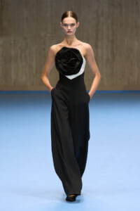 Model walking a black strapless jumpsuit with a large black fabric rose on the chest, white panel at the bodice, on a blue runway.