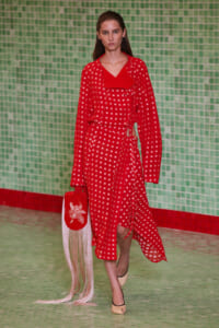 Model on a runway wearing a red patterned dress with a waist tie, carrying a red fringe bag, against a green tiled backdrop.