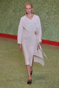 Model walks the runway in an oversized white wrap coat with tied belts, against a green tiled wall.