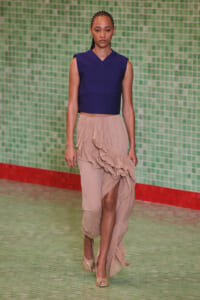 Model walking a runway in a navy sleeveless crop top and beige tiered ruffled skirt against a green tiled backdrop.