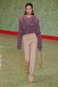 Model on runway wearing a purple ruched, ruffled blouse and beige textured pants with a tan belt, carrying a beige handbag.