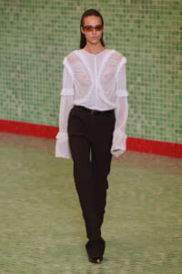 Model on a runway wearing a sheer white ruffled blouse with long sleeves and black textured pants, brown sunglasses, against a green tiled wall.
