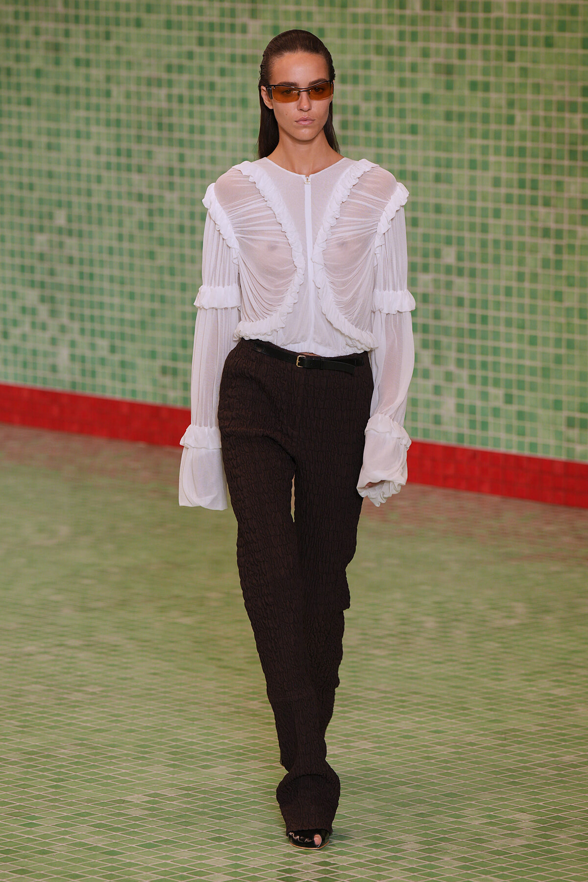 Model on a runway wearing a sheer white ruffled blouse with long sleeves and black textured pants, brown sunglasses, against a green tiled wall.