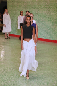 Model walks the runway in a sleeveless black cropped top and a white layered flowing skirt, beige peep-toe heels, against a green tiled backdrop.