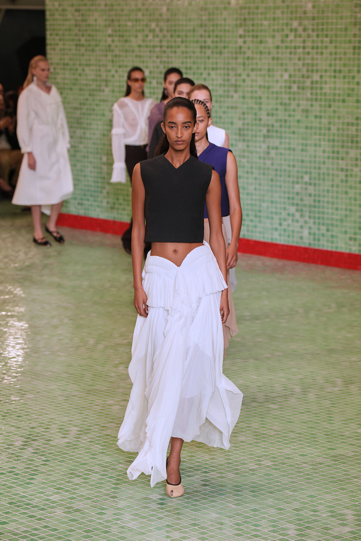 Model walks the runway in a sleeveless black cropped top and a white layered flowing skirt, beige peep-toe heels, against a green tiled backdrop.