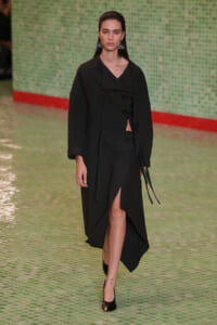 Model wearing an all-black, long coat with a high slit and tied closures, walking on a green tiled runway with a red edge in the background.