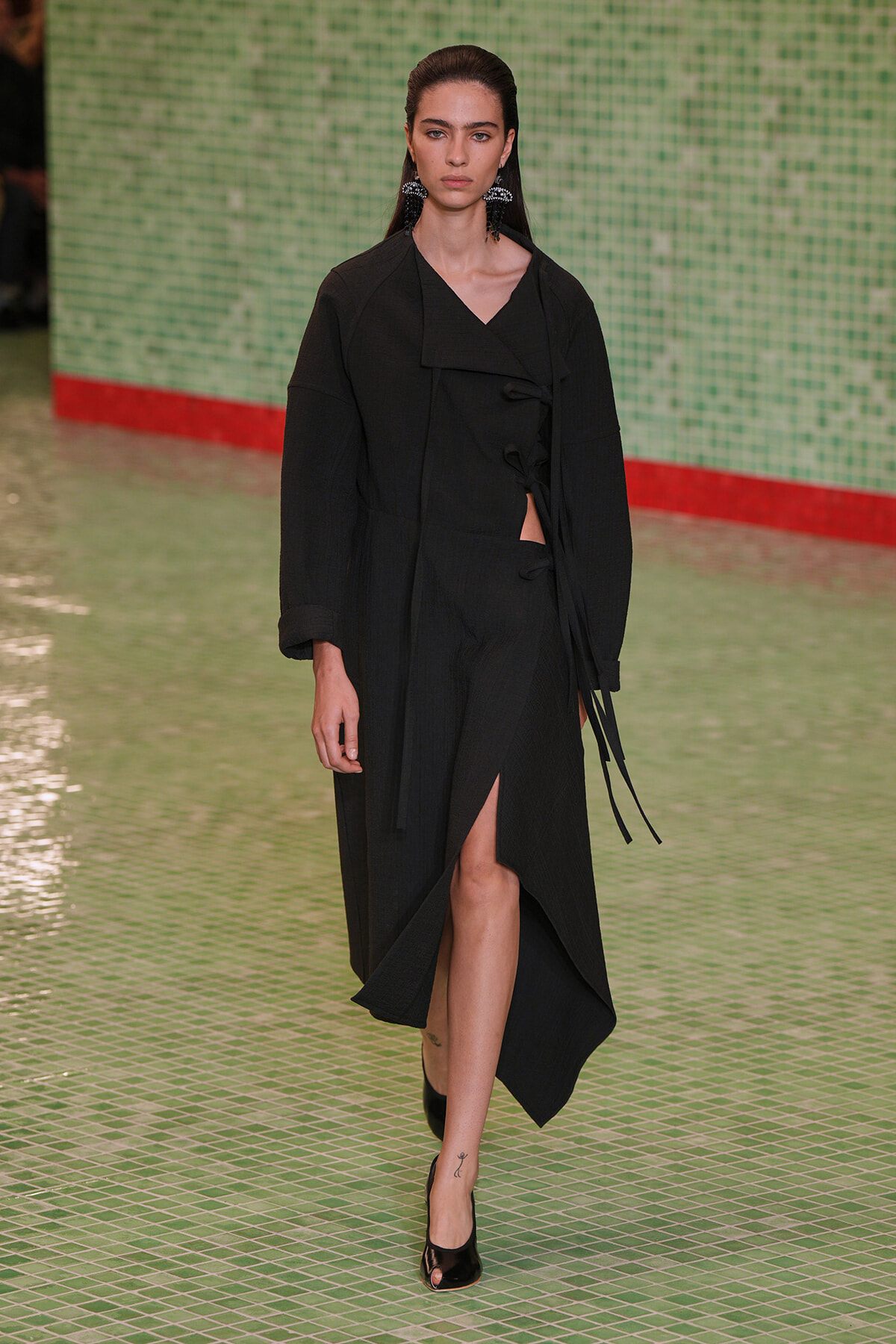 Model wearing an all-black, long coat with a high slit and tied closures, walking on a green tiled runway with a red edge in the background.
