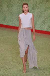 Model walks the runway in a white sleeveless crop top and a pale gray layered asymmetrical skirt with a high slit, beige heels.