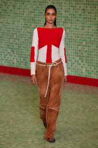 Model walking the runway in a red-and-white color-block knit sweater, brown suede wide-leg pants with a white rope belt, and yellow-tinted glasses against a green tiled backdrop.