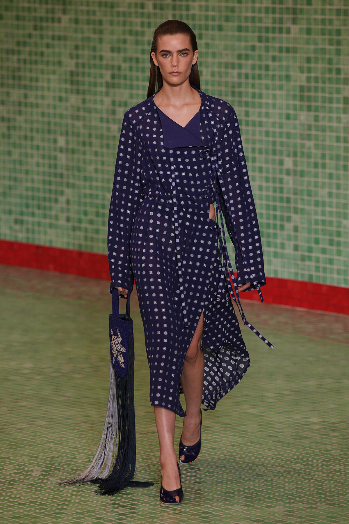 Model walking runway in a navy patterned dress with white squares, wrap belt, and side slit in a green tiled backdrop.