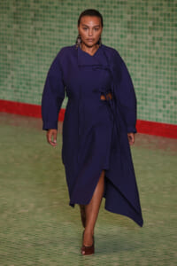 Model walking runway in a navy blue wrap coat with waist ties and an asymmetric hem, paired with brown heels.