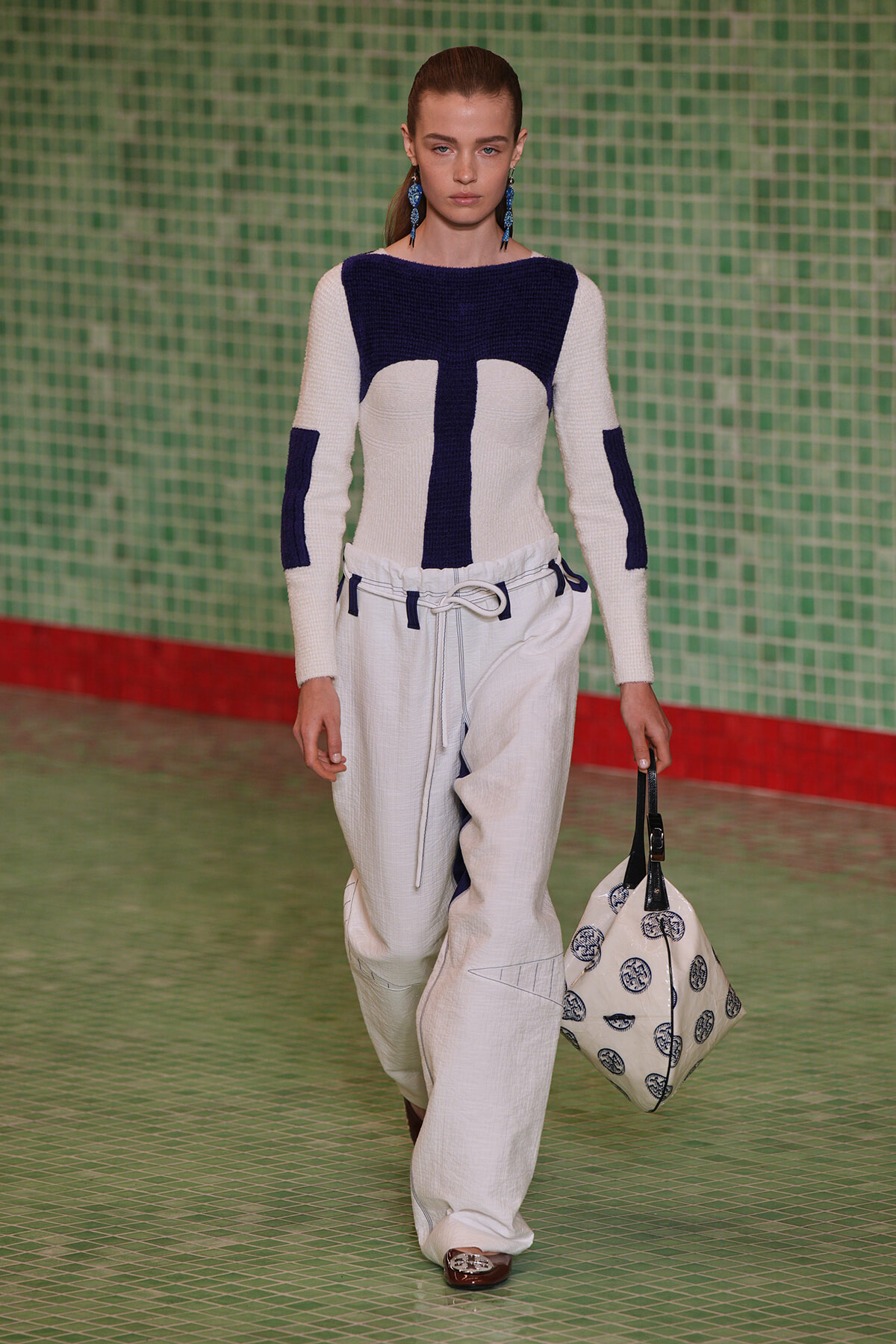 Model walking the runway in a white and navy color-block knit sweater paired with wide white pants and a white rope belt, carrying a patterned tote bag.