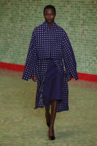 Model walking a runway in a navy blue coat with white square patterns over a matching outfit, against a green tiled backdrop.