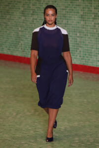 Model walking a runway in a navy blue dress with white and black color-block sleeves, teal-green tiled backdrop.