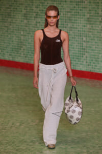 Model walks a runway in a brown sequined tank top, white striped wide-leg pants with a rope belt, tan shoes, and a white patterned tote bag against a green tiled backdrop.