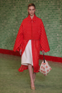 Runway model in a red coat with small white square pattern over a white dress, beige heels, and a white patterned bag against a green tiled backdrop.