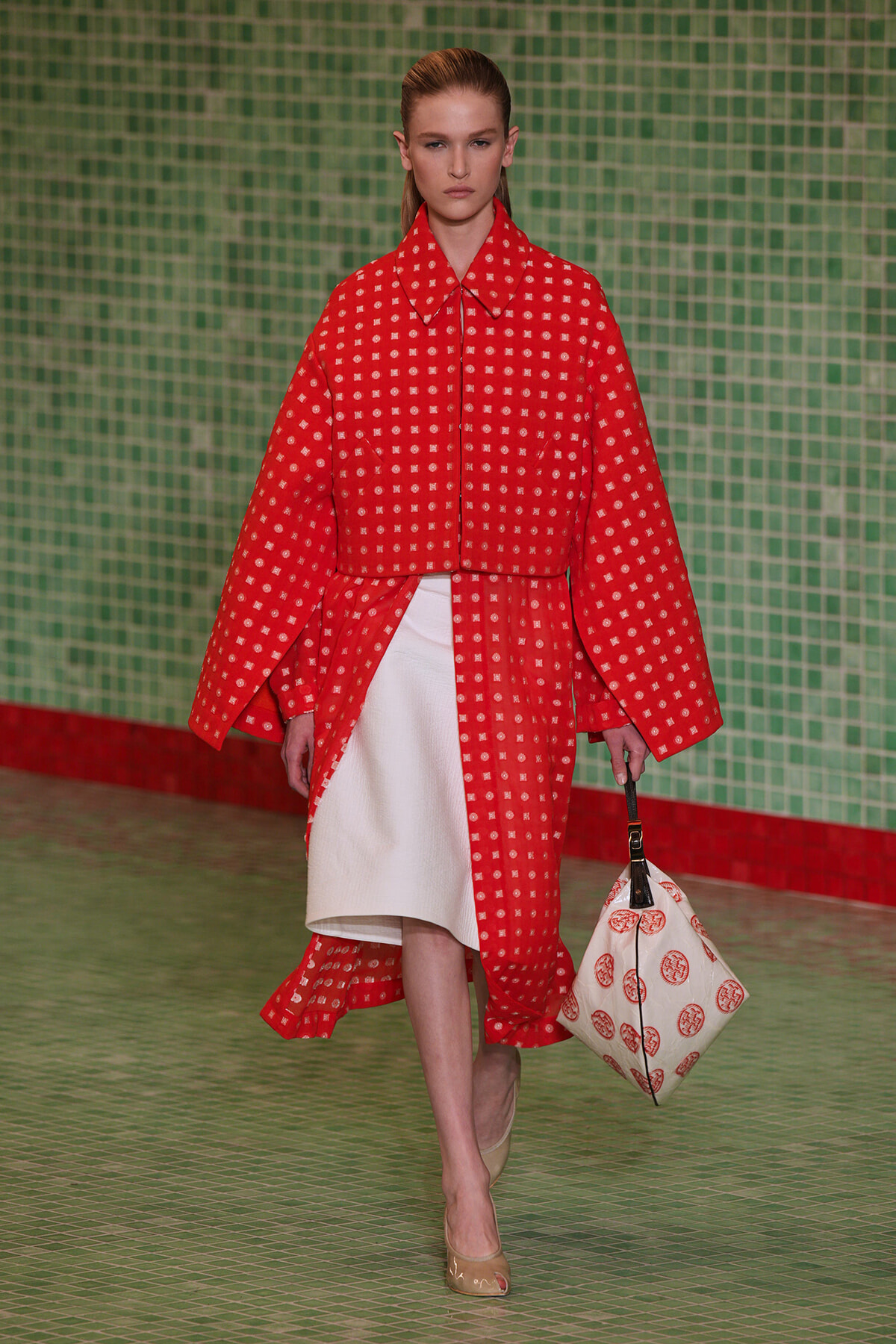 Runway model in a red coat with small white square pattern over a white dress, beige heels, and a white patterned bag against a green tiled backdrop.