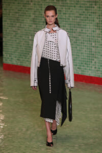 Model walking a runway in a white jacket with black piping over a white printed top and a black layered skirt, carrying a black handbag.