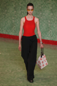 Model walking a runway in a red textured tank top and black wide-leg pants, holding a white tote with red circular prints and wearing yellow sunglasses.