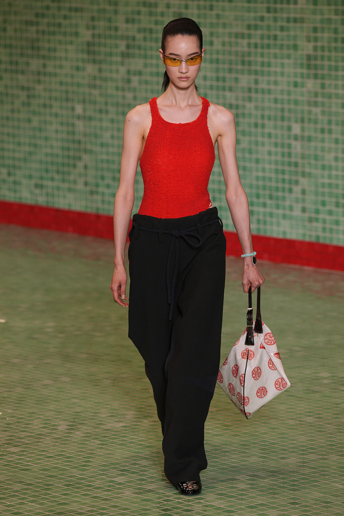 Model walking a runway in a red textured tank top and black wide-leg pants, holding a white tote with red circular prints and wearing yellow sunglasses.