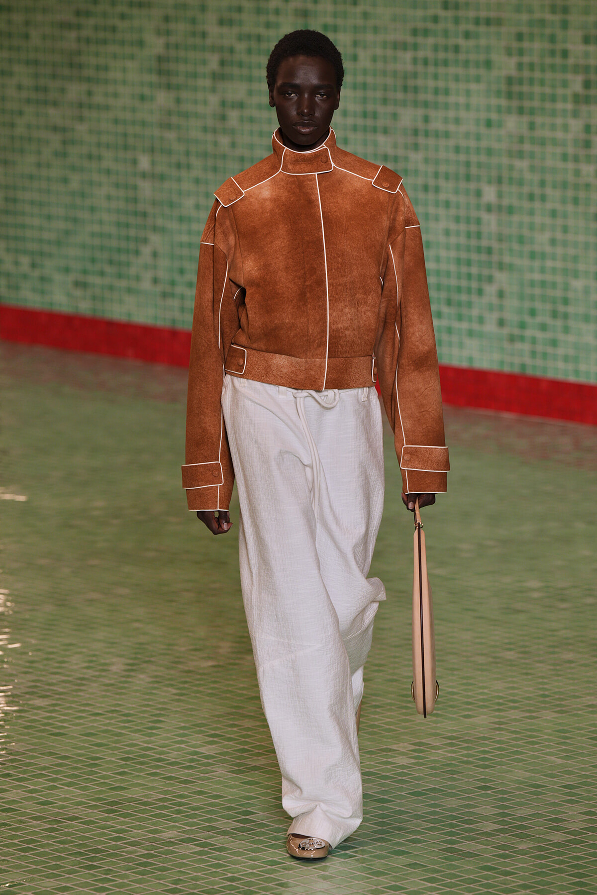 Model wearing a brown suede jacket with white piping and wide beige trousers, carrying a beige bag on a green-tiled runway setting.
