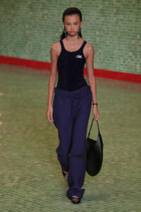 Model walking runway in a navy sequined tank top and wide-leg blue pants, carrying a black shoulder bag in a fashion show.