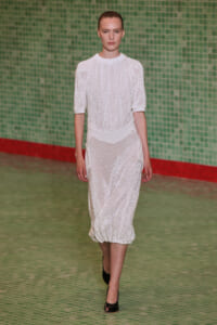 Model walking a runway in a white textured knit midi dress with short sleeves against a green tiled wall.