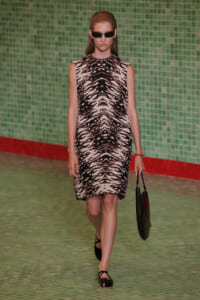 Model on a runway wearing a sleeveless beige and black tiger‑print dress, black sunglasses, and carrying a black handbag.