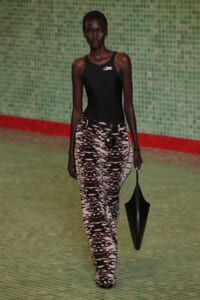 Runway model with dark skin wearing a black sleeveless top and zebra-print tan-and-black wide-leg pants, a black belt with a round accessory, and holding a black umbrella-shaped bag against a green tiled backdrop.