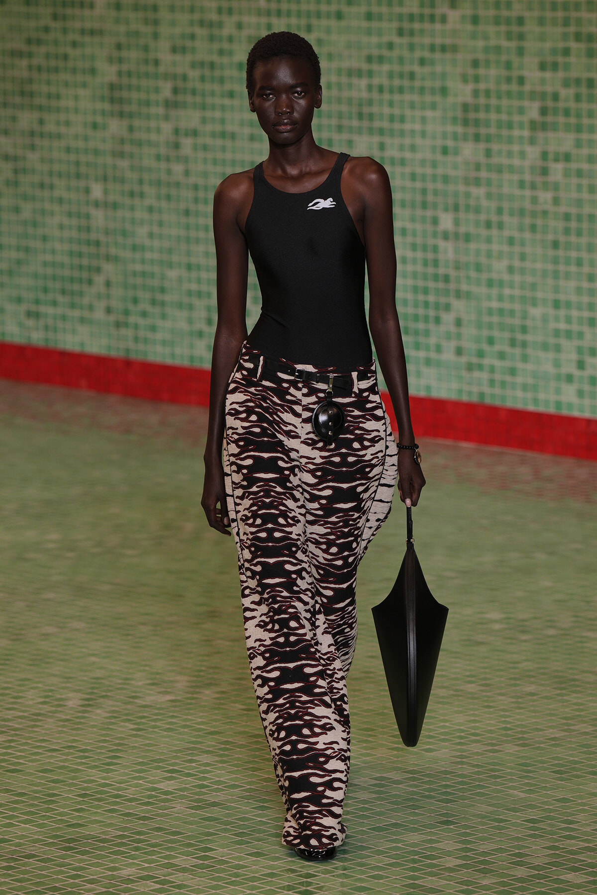 Runway model with dark skin wearing a black sleeveless top and zebra-print tan-and-black wide-leg pants, a black belt with a round accessory, and holding a black umbrella-shaped bag against a green tiled backdrop.