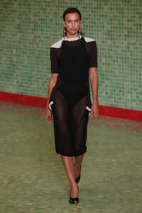 Model walks a runway in a sheer black dress with white shoulder panels and black high heels.