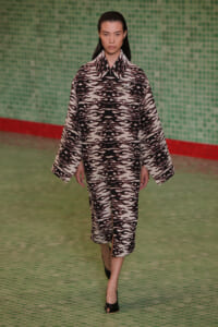 Model walking runway in a black-and-white flame-pattern matching coat and skirt set, green tiled backdrop behind her.