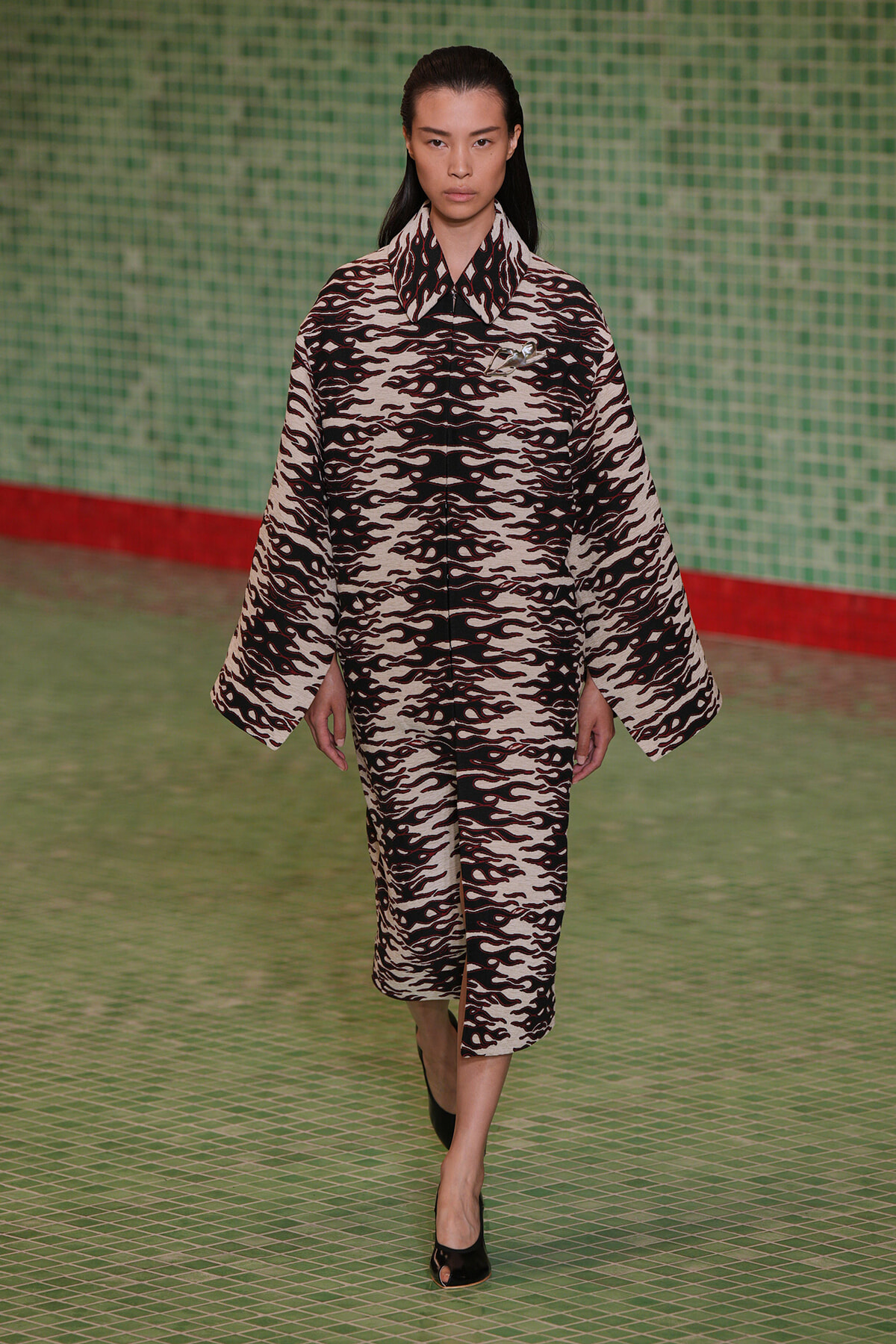 Model walking runway in a black-and-white flame-pattern matching coat and skirt set, green tiled backdrop behind her.